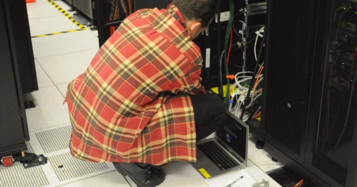 A technician crouched beside an open server rack, working on a laptop on the floor of a data centre
